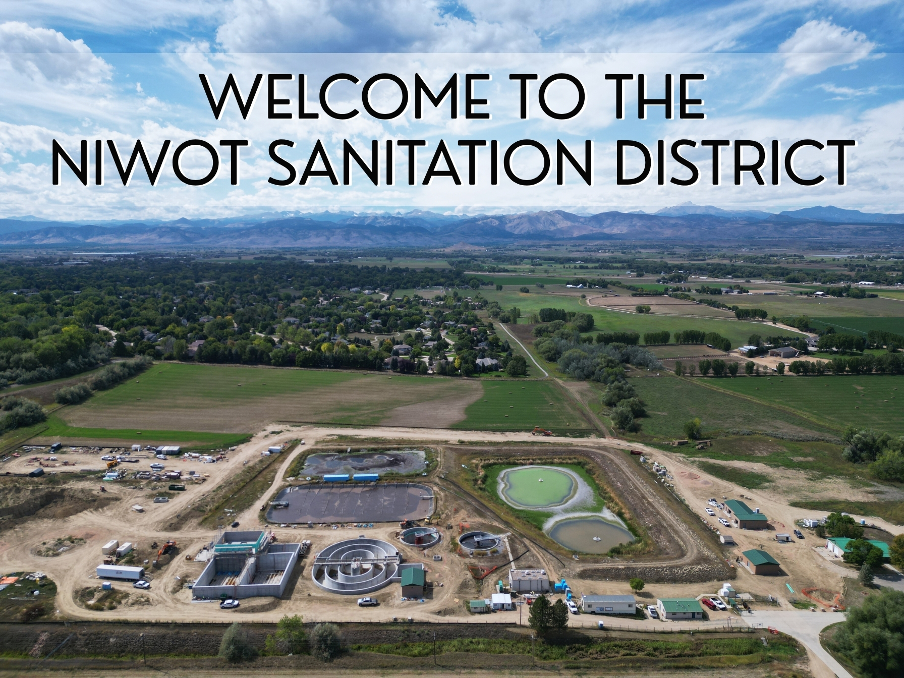 Aerial view of the Niwot Sanitation District facility with a "Welcome to the Niwot Sanitation District" sign overlay. The image shows a sprawling wastewater treatment plant with various structures, ponds, and construction areas, surrounded by green fields, residential neighborhoods, and distant mountains under a partly cloudy sky.