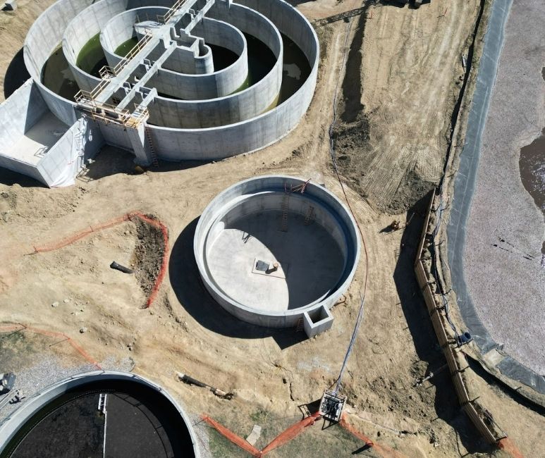Aerial view of a construction site at Niwot Sanitation, featuring a large circular Oxidation Ditch with green water undergoing water testing. The ditch has concentric sections with walkways and scaffolding. A smaller circular tank and a rectangular structure with set forms for the Was and Ras building are nearby, surrounded by a dirt area with construction materials, temporary fencing, and a lined pond in the background.