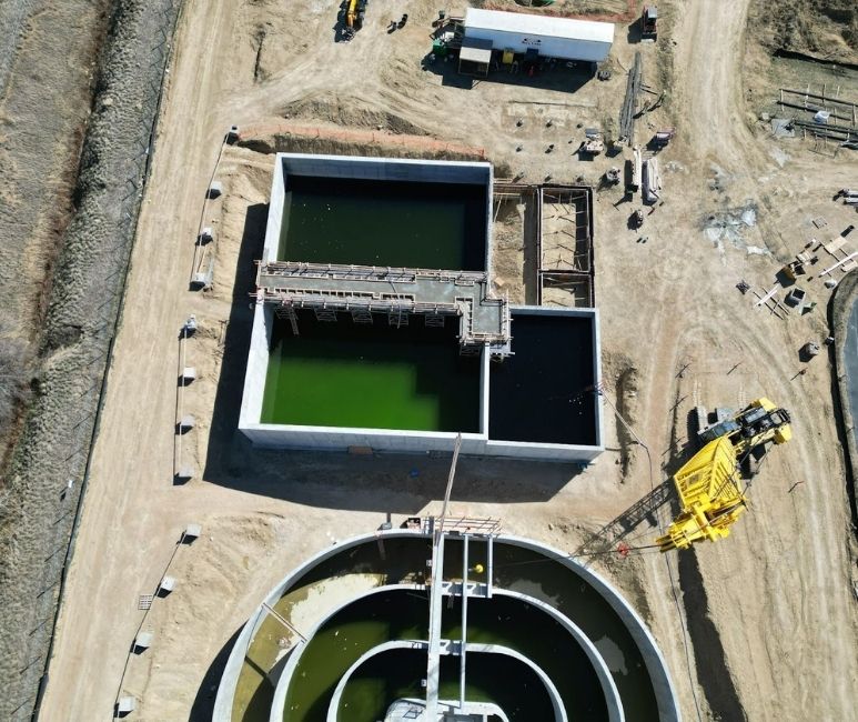 Aerial view of a construction site at Niwot Sanitation District, showing large concrete tanks with green water, part of a wastewater treatment facility. A yellow crane is positioned near a circular tank, while rectangular tanks with scaffolding and walkways are under construction. Construction materials, a white trailer, and heavy machinery are scattered around the site, with temporary fencing and a dirt path visible in the background.