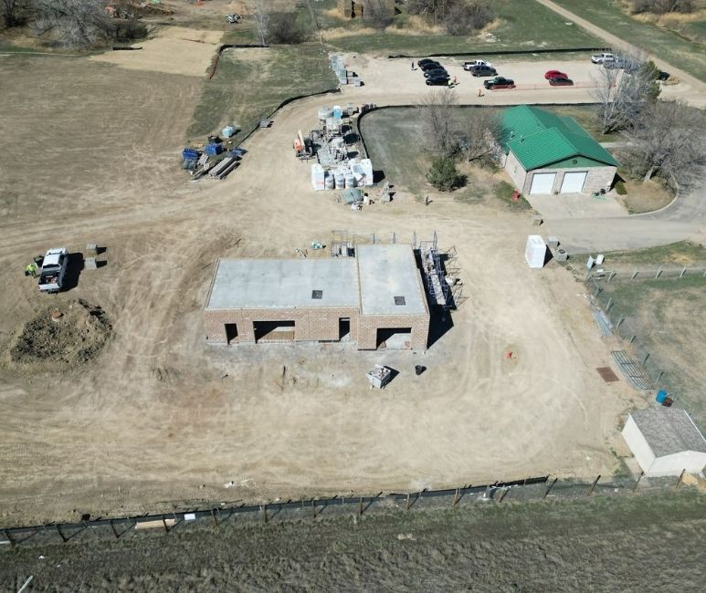 Aerial view of a construction site at Niwot Sanitation District, showcasing the Headworks building with CMU walls installed and a recently poured roof slab, awaiting trusses and roofing. The brick structure is surrounded by a fenced area with construction materials, a white pickup truck, and scattered equipment. Nearby are a green-roofed shed, temporary structures, and a parking area with several vehicles. A dirt path and grassy terrain are visible in the background.