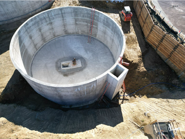 Aerial view of a large, circular concrete tank under construction. The tank has a central opening with a small concrete structure in the middle. A ladder leans against the side of the tank. A small excavator is working on the side of the tank.