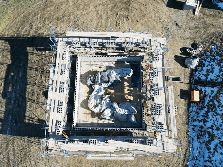 Aerial view of the Contact Chamber construction site, showing a rectangular structure outlined with CMU blocks and surrounded by scaffolding. Inside the structure, tarped materials are visible, with patches of snow and dirt around the site.