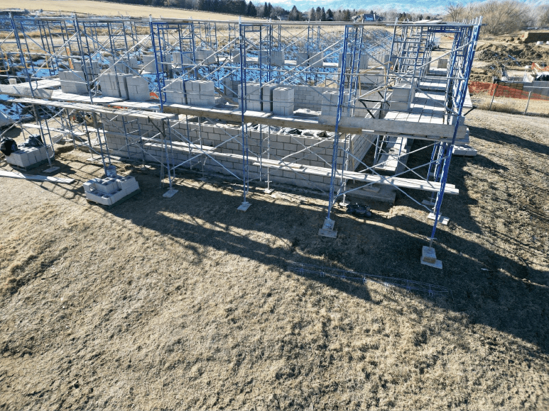 Ground-level view of the Contact Chamber construction site, showing scaffolding surrounding a partially built structure made of CMU blocks. Stacks of unused CMU blocks and construction materials are visible on the ground, with an open field in the background.