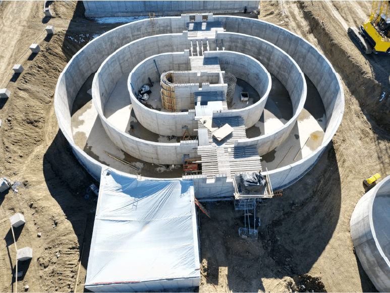 Aerial view of a large circular concrete structure under construction. The structure has multiple concentric rings and a central core.