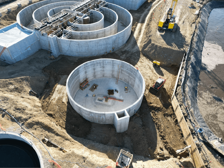 An aerial view of a construction site featuring a large, circular concrete structure with a smaller square section at its center. Surrounding the main structure are several curved concrete walls forming a spiral pattern. Construction equipment and workers are visible on site. The area is bordered by dirt paths and temporary fencing, with a crane and other machinery positioned nearby.