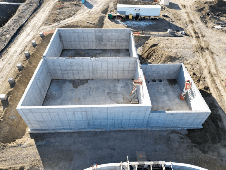 An aerial view of a construction site showing a large concrete foundation with two rectangular sections. The site is surrounded by dirt roads and construction materials, with some equipment and a white truck with 'Rice Lake' branding in the background.