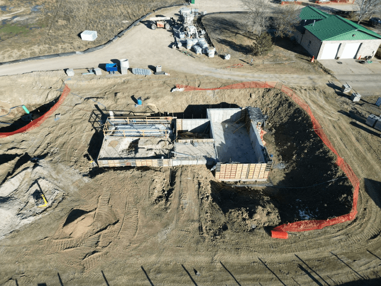 Aerial view of a construction site with an excavated area showing the foundation of a building. The site is surrounded by construction materials, equipment, and a green-roofed building nearby. Safety fencing in red and black lines the perimeter of the excavation.