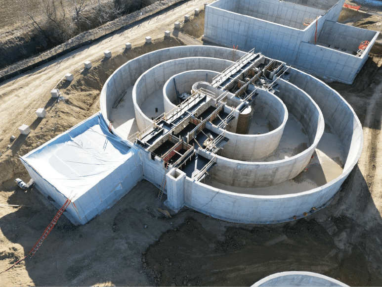 Aerial view of a construction site featuring an oxidation ditch with multiple concentric concrete rings and elevated steel frameworks for future concrete pours. Adjacent structures are also under construction, with some areas covered by protective white sheeting, and a dirt road running alongside. The site is surrounded by barren land with sparse vegetation.