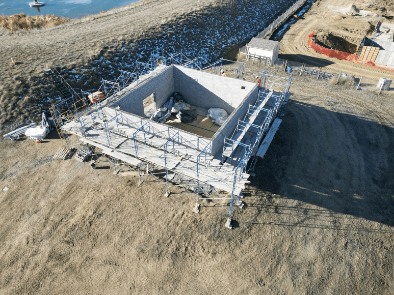 An aerial view of a construction site where the UV building is taking shape. The structure is surrounded by scaffolding, with workers visible on site. The building's concrete block walls are partially erected, and the interior contains construction materials and equipment. The site is bordered by a chain-link fence and is situated near a body of water, with some snow patches visible on the ground.