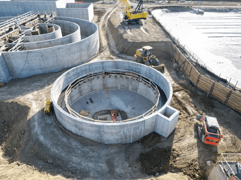 An aerial view of a large construction site featuring a circular concrete clarifier under construction. The site includes heavy machinery such as a yellow loader and a crane, with visible construction materials and temporary fencing around the area. The surrounding area is in various stages of development with dirt paths and other construction activities.