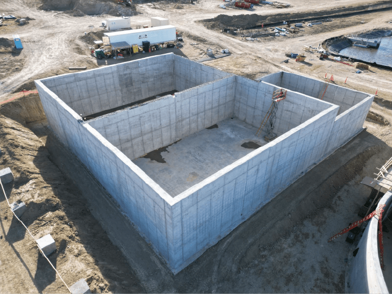 An aerial view of a large, rectangular concrete structure under construction, identified as a dewatering building. The site shows scaffolding and construction equipment, with surrounding dirt paths and other construction activities in the background.