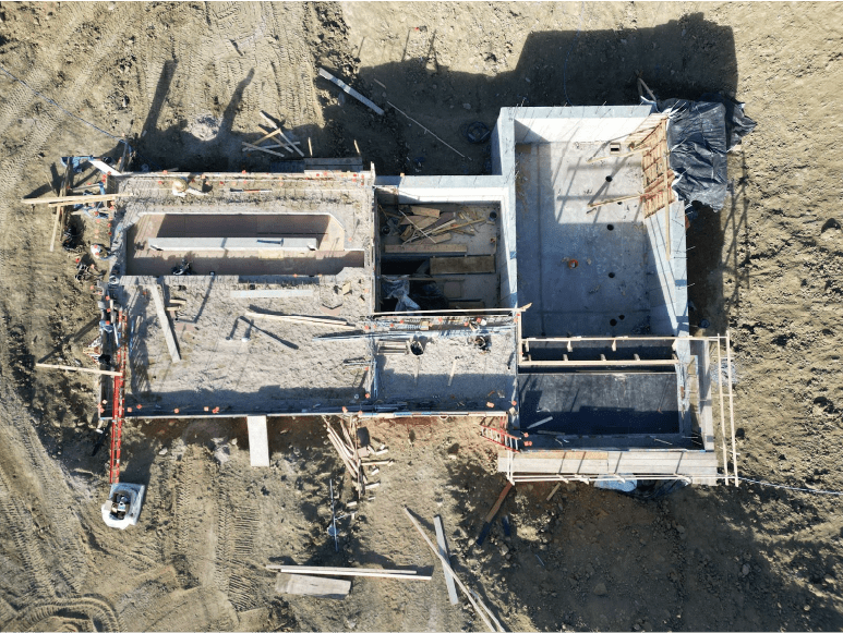 An aerial view of a construction site showing a partially built structure with concrete walls and floors. There are scattered construction materials, wooden planks, and workers on site. The area around the structure is mostly dirt with some construction equipment visible.