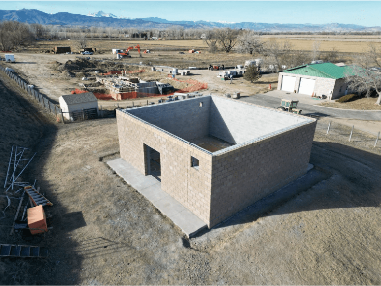 An aerial view of a construction site in a rural area with mountains in the background. The main structure is a partially completed square building made of concrete blocks with a doorway and small window. Construction materials and equipment are scattered around, including a pile of metal rods and an orange box nearby. In the background, there's a green-roofed building and various construction machinery and workers. The site is surrounded by open fields and sparse trees.