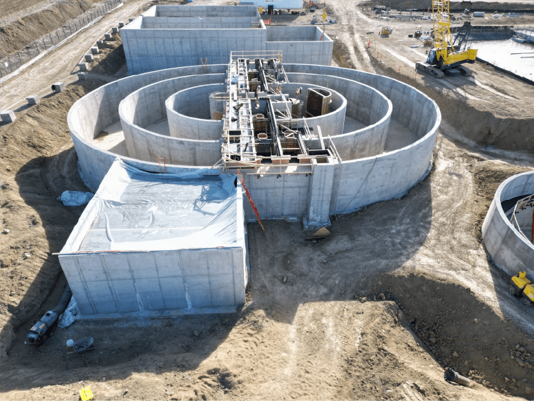 Construction site of an Oxidation Ditch, featuring multiple circular concrete structures and scaffolding for elevated decks. Workers and machinery are present, with a yellow crane and construction materials scattered around the site. The area is surrounded by dirt paths and fencing.