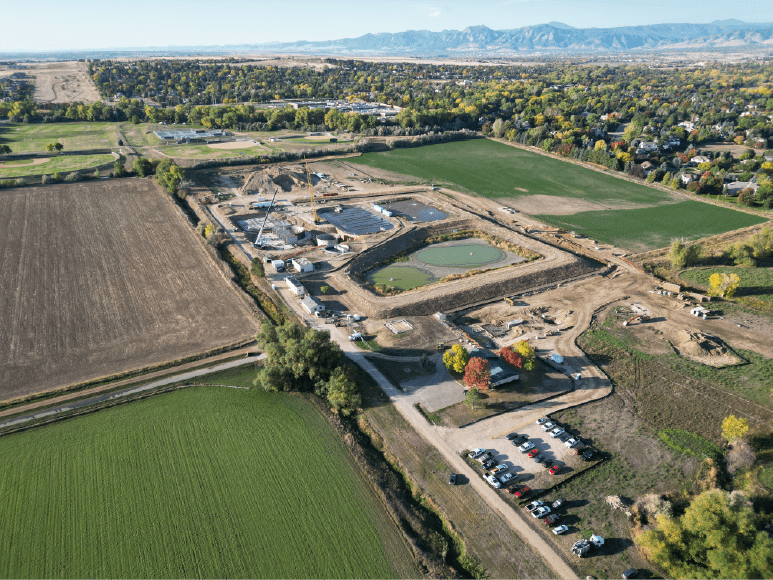 An aerial view of a construction site surrounded by agricultural fields and greenery. The site includes various buildings under construction, visible cranes, excavation work, and water bodies within the boundaries. A parking area with numerous vehicles is located near the bottom, and in the background, there are mountains and a mix of residential and recreational areas with open fields and trees in vibrant autumn colors.