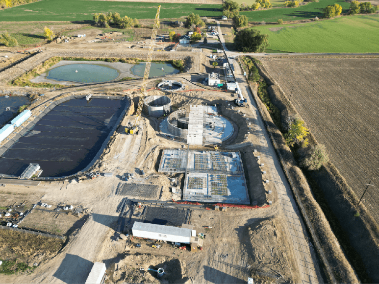 Aerial view of a large construction site at a wastewater treatment plant. There are several circular structures in various stages of construction, as well as large rectangular tanks and ponds. Heavy machinery, such as cranes and excavators, are visible on the site. The surrounding area is rural farmland.