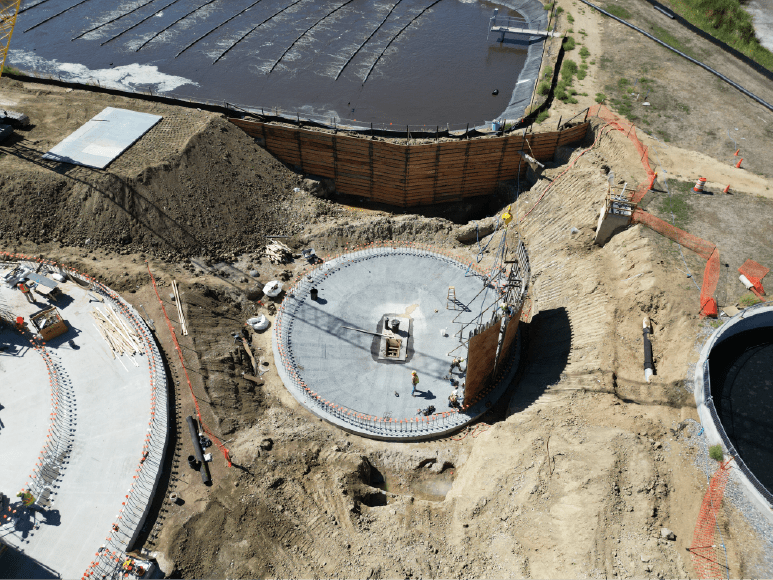 Aerial view of a construction site showing a circular foundation with wooden formwork in place. The foundation has a central concrete slab with metal rebar protruding. There are several other smaller structures and equipment scattered around the site.