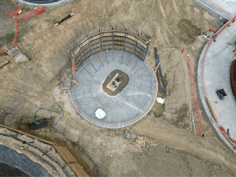 Aerial view of a construction site showing a circular foundation with wooden formwork in place. The foundation has a central concrete slab with metal rebar protruding. There are several other smaller structures and equipment scattered around the site.