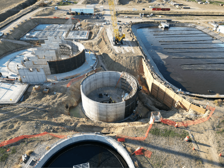 Aerial view of a large construction site with multiple circular structures in various stages of completion. The structures have wooden formwork and metal rebar, and there are several pieces of heavy machinery on site. The surrounding area is a mix of farmland and industrial buildings. 