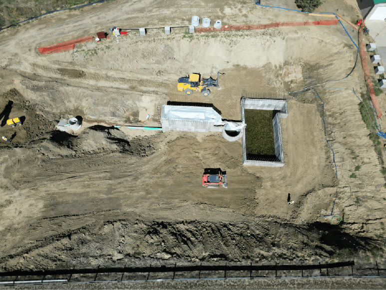 Aerial view of a construction site with heavy machinery, including a backhoe and a bulldozer. There is a large rectangular pit with concrete walls being built, and several other smaller structures and pipes are visible. The surrounding area is mostly dirt and gravel. 