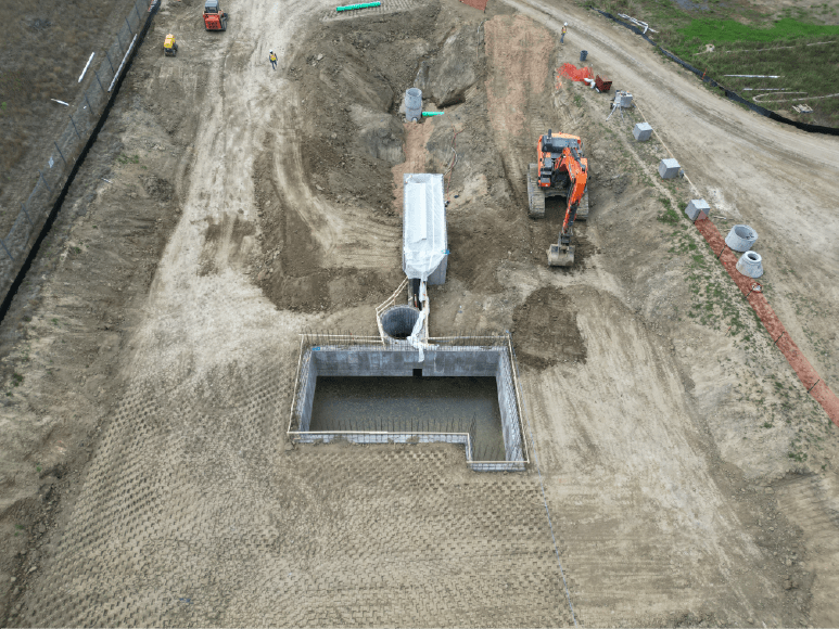 Aerial view of a construction site with heavy machinery, including an excavator. There is a large rectangular pit with concrete walls being built, and several other smaller structures and pipes are visible. The surrounding area is mostly dirt and gravel. 