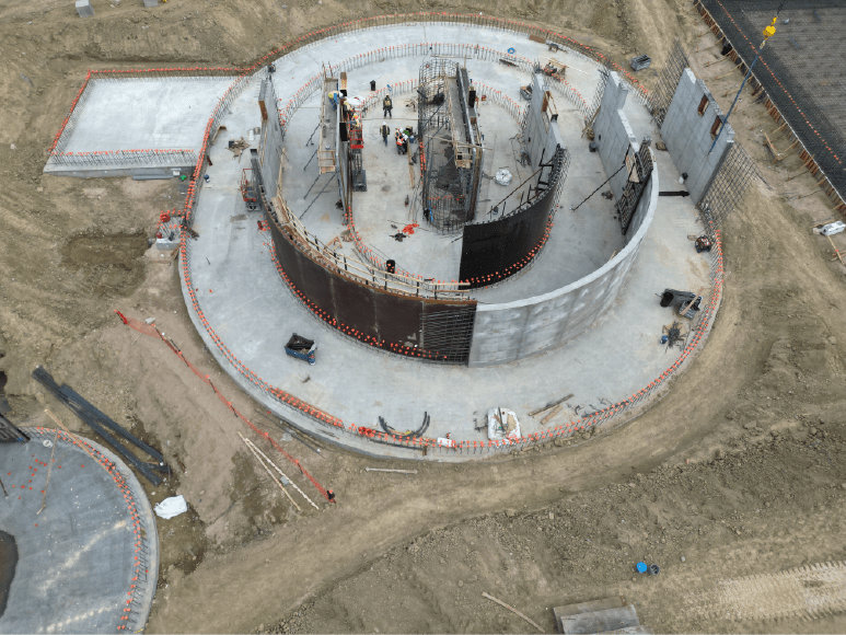 An overhead view of a circular concrete structure under construction, likely part of the Oxidation Ditch. The inner walls are being formed and reinforced with rebar, with several workers actively engaged in the process. The smooth concrete foundation is already in place, and scaffolding and construction materials are visible around the site. The surrounding area shows dirt paths and construction activity, indicating the ongoing development of this critical structure.