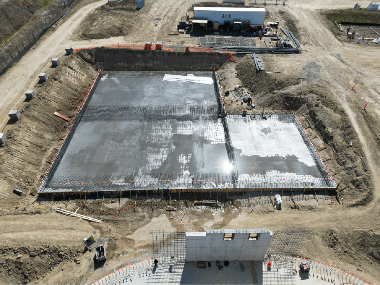 An overhead view of a freshly poured concrete slab at a construction site, still wet from the curing process. The rectangular slab is surrounded by rebar and earthworks, with visible construction materials and equipment nearby. The curing water gives the slab a reflective sheen, while workers and machinery are positioned around the site, indicating active construction progress. Dirt paths and surrounding structures outline the site's boundaries.