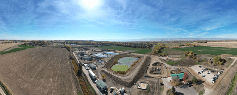 A panoramic aerial photograph of a large water treatment facility under construction. The facility is located in a rural area with fields and trees visible in the distance. Several large concrete structures and pools of water can be seen within the facility. Construction equipment, including trucks and cranes, is visible on the site.