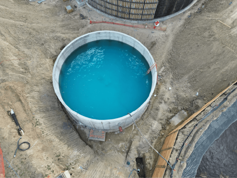 Aerial view of a clarifier filled with bright blue water, marking the start of a water test. The circular concrete structure is surrounded by dirt and excavation, with equipment and safety barriers nearby. This test provides a rare look at the clarifier basin before final backfill and further construction work, such as the installation of the scum pit.