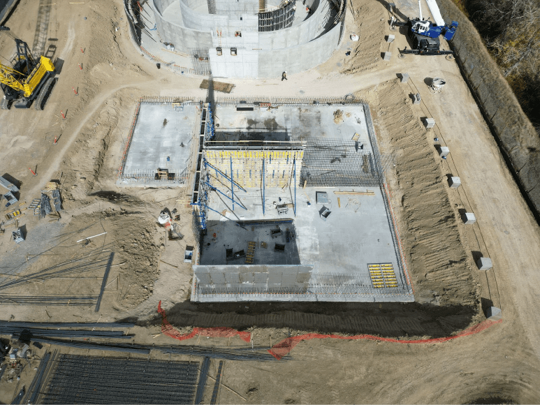 An aerial photograph of a large construction site showing a partially built concrete structure. The structure has a large, curved wall and a smaller, rectangular section. Several smaller concrete structures and piles of dirt are visible around the main structure. Construction equipment, including cranes, is also visible on the site.