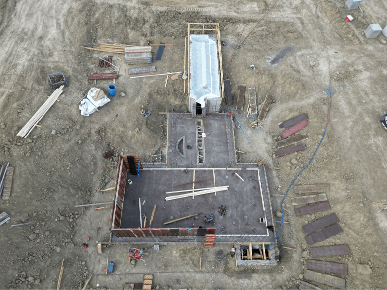 Aerial view of a construction site with concrete recently poured in the headworks area. Several wall forms are installed, with some tools and materials scattered around the site. The covered structure remains in place, while construction equipment and wooden planks are visible, indicating ongoing work. The area around the site is mostly cleared, with a few construction materials and panels on the ground.