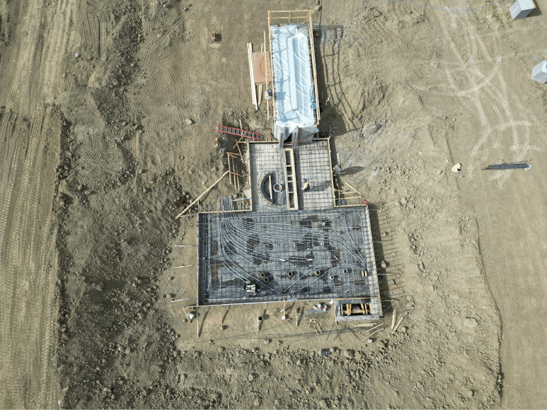 An aerial view of the construction site, showing significant progress at the headworks. Rebar is fully installed across a large grid-like section, with additional formwork in place for walls. A covered structure sits at the upper part of the site, indicating areas prepared for concrete pours or protection from elements. The surrounding area is cleared, with visible tracks and some equipment on site.