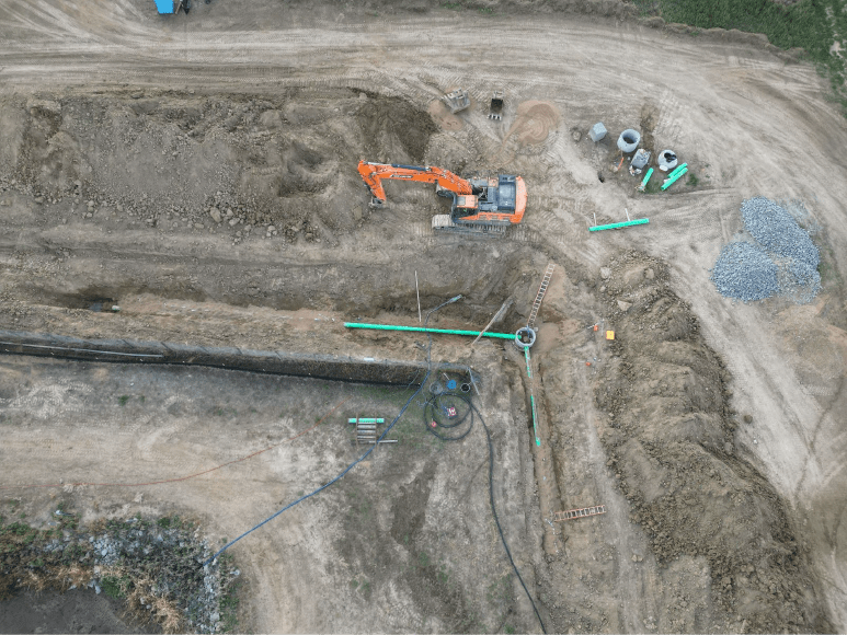 An aerial photograph of a construction site with a large orange excavator digging a trench in the dirt. The trench is long and narrow, and there are several pipes and other construction materials visible along the sides of the trench. A pile of rocks and a few smaller pipes are visible in the top right corner of the image.