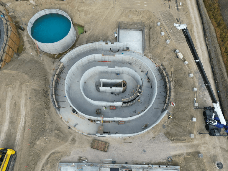 An aerial photograph of a large, circular concrete structure in the middle of a construction site. The structure appears to be a water treatment facility, with a central circular opening and concentric rings forming a basin. To the left of the structure, there is a large, circular pool filled with blue water. To the right, there is a smaller, rectangular building with a flat roof. Construction equipment, including cranes and excavators, is visible on the site.