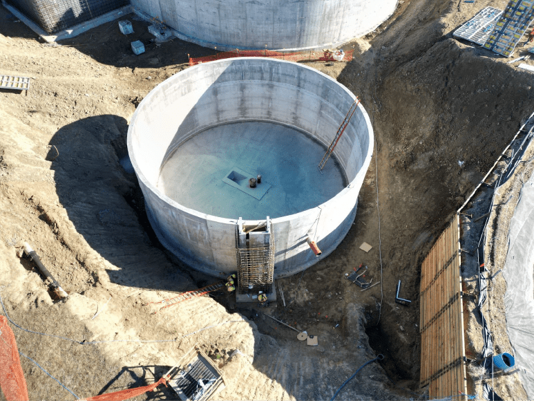 Aerial view of a large, circular concrete tank under construction. The tank is surrounded by dirt and excavation equipment. Workers are visible on the site.