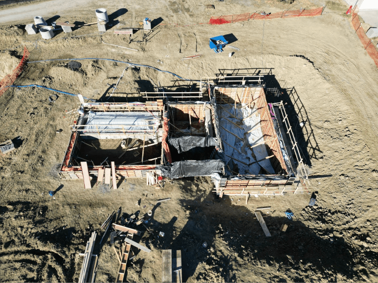 Aerial view of the Headworks project construction site, showing completed wall pours covered with blankets and surrounded by a structured framework. The area is secured with safety barriers, and construction materials are visible on the ground nearby.