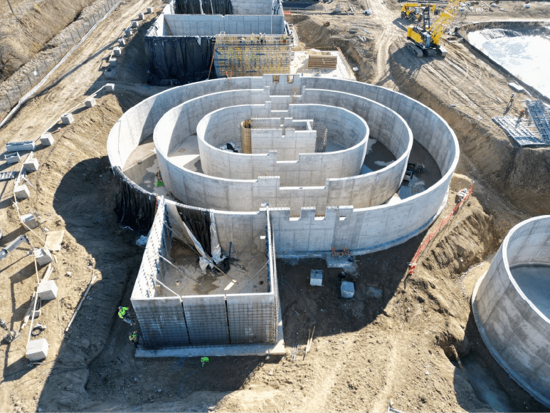 An aerial view of the construction site showing the Oxidation Ditch anaerobic tank taking shape, featuring circular concrete walls, rebar installations, and surrounding construction activity.