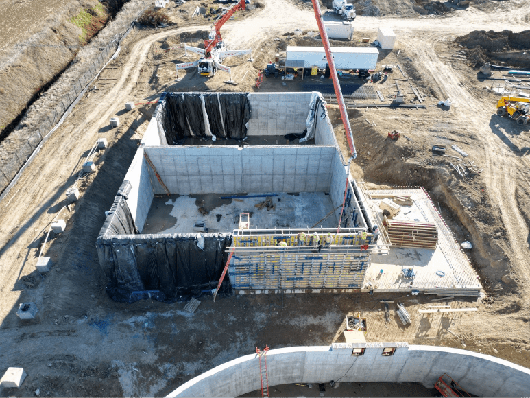 A construction site for the Dewatering building, showcasing significant progress. The aerobic digester tanks are enclosed within concrete walls, with black tarps draped over portions to retain heat during the curing process. Construction workers and equipment are visible, along with formwork and materials being prepared for additional building phases.