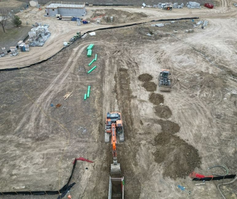 Aerial view of a construction site at Niwot Sanitation, showing an orange excavator backfilling a trench near green pipes. The site includes various construction materials, a white pickup truck, temporary fencing, and heavy machinery scattered around. A concrete building under construction and a dirt path are visible, with a grassy landscape in the background.