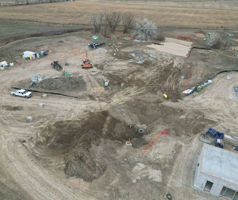 Aerial view of a construction site at Niwot Sanitation, showing an orange excavator backfilling a trench near green pipes. The site includes various construction materials, a white pickup truck, temporary fencing, and heavy machinery scattered around. A concrete building under construction and a dirt path are visible, with a grassy landscape in the background.