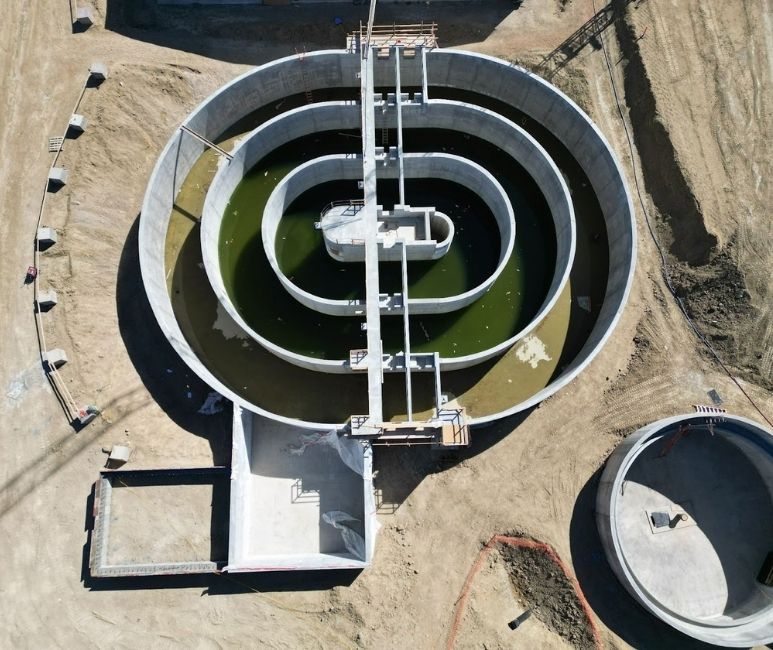 Aerial view of a construction site at Niwot Sanitation, featuring a large circular concrete tank with green water, part of a wastewater treatment facility. The tank has concentric sections and a central structure with walkways and scaffolding. A smaller circular tank and a rectangular concrete structure are nearby, surrounded by a dirt area with construction materials and temporary fencing.