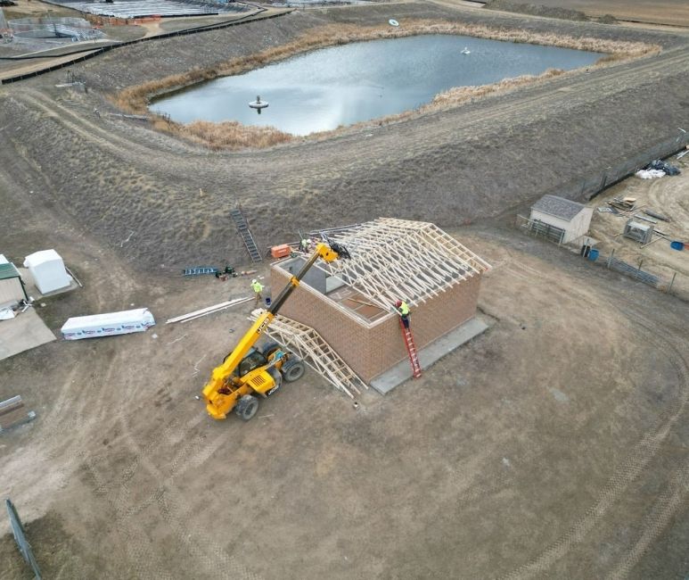 Aerial view of a construction site near a pond. A brick building with a partially completed wooden roof is in the foreground, with two workers on the roof and a yellow telehandler nearby. Construction materials and a trailer are scattered around the site. The pond in the background has a fountain and a small boat on its surface, surrounded by a dirt embankment.