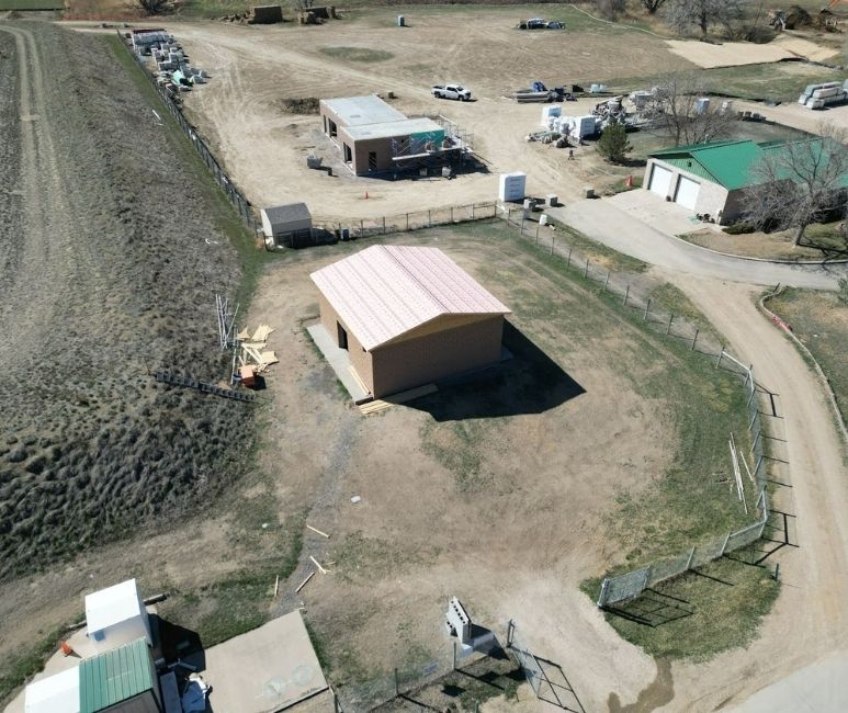 Aerial view of a construction site at Niwot Sanitation District, featuring a UV building with trusses and weather guard installed, awaiting metal sheeting. The brick structure is surrounded by a fenced area with scattered construction materials, a dirt path, and nearby buildings, including a green-roofed shed and temporary structures. A small excavator and stacks of supplies are visible in the background.