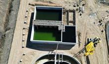 Aerial view of a construction site at Niwot Sanitation District, showing large concrete tanks with green water, part of a wastewater treatment facility. A yellow crane is positioned near a circular tank, while rectangular tanks with scaffolding and walkways are under construction. Construction materials, a white trailer, and heavy machinery are scattered around the site, with temporary fencing and a dirt path visible in the background.