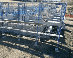 Ground-level view of the Contact Chamber construction site, showing scaffolding surrounding a partially built structure made of CMU blocks. Stacks of unused CMU blocks and construction materials are visible on the ground, with an open field in the background.