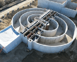 Aerial view of a construction site featuring an oxidation ditch with multiple concentric concrete rings and elevated steel frameworks for future concrete pours. Adjacent structures are also under construction, with some areas covered by protective white sheeting, and a dirt road running alongside. The site is surrounded by barren land with sparse vegetation.