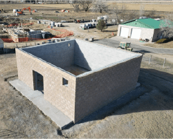 An aerial view of a construction site in a rural area with mountains in the background. The main structure is a partially completed square building made of concrete blocks with a doorway and small window. Construction materials and equipment are scattered around, including a pile of metal rods and an orange box nearby. In the background, there's a green-roofed building and various construction machinery and workers. The site is surrounded by open fields and sparse trees.