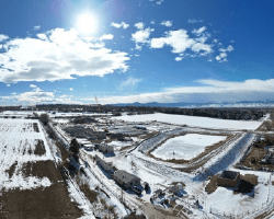 A panoramic aerial view of a rural construction site under a bright blue sky with scattered clouds. The site is surrounded by snow-covered fields, with patches of bare ground visible. In the foreground, there are several buildings and structures, including what appears to be a construction office and storage facilities. Construction equipment and vehicles are scattered around, with dirt roads and pathways connecting different parts of the site. In the background, the landscape extends into flat, open fields