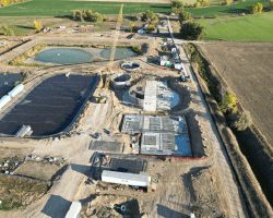 Aerial view of a large construction site at a wastewater treatment plant. There are several circular structures in various stages of construction, as well as large rectangular tanks and ponds. Heavy machinery, such as cranes and excavators, are visible on the site. The surrounding area is rural farmland.