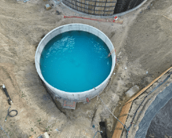 Aerial view of a clarifier filled with bright blue water, marking the start of a water test. The circular concrete structure is surrounded by dirt and excavation, with equipment and safety barriers nearby. This test provides a rare look at the clarifier basin before final backfill and further construction work, such as the installation of the scum pit.